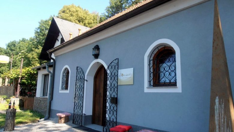 Mal&aacute; modr&aacute; budova s dřevěn&yacute;mi dveřmi a ozdobn&yacute;mi mř&iacute;žemi před okny. Před n&iacute; stoj&iacute; dřevěn&eacute; stoličky s červen&yacute;mi pol&scaron;t&aacute;ři.