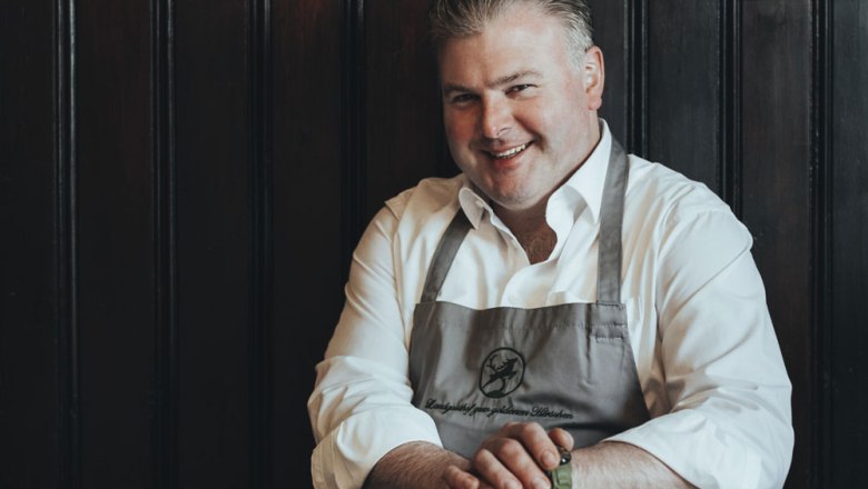 Man in a white shirt and apron smiles in front of a dark wooden wall.