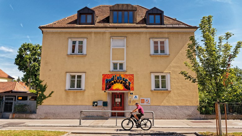 Yellow building with red entrance and graffiti sign. A cyclist rides past. Blue sky and trees in the background.
