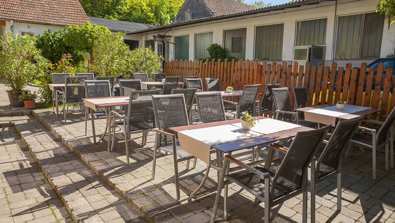 Gastgarten mit Tischen und Stühlen im Freien, umgeben von Pflanzen und einem Holzzaun.