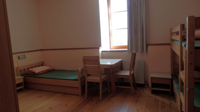 Ein einfach eingerichtetes Gästezimmer mit Holzboden, Einzelbett, Etagenbett, Tisch und Stühlen, sowie einem Fenster mit Vorhang.