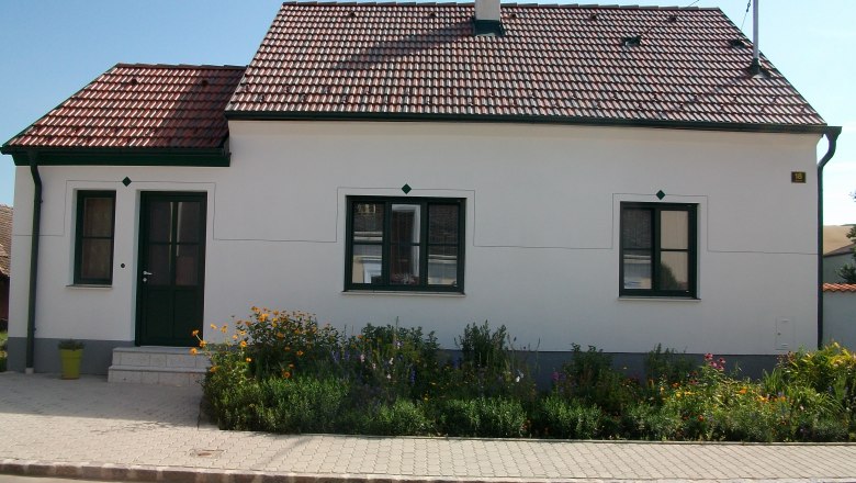 A small, white house with a red tiled roof and green door frame, surrounded by a well-tended garden with flowers.