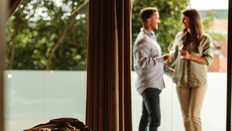 Vřelé přivítání, © Weinviertel Tourismus GmbH / Michael Reidinger Pár stojí na balkoně a cinká skleničkami, zatímco v popředí leží na posteli cestovní taška.