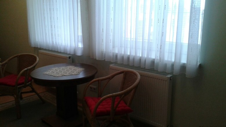 Zwei Korbst&uuml;hle mit roten Kissen und ein runder Holztisch vor einem Fenster mit wei&szlig;en Vorh&auml;ngen.