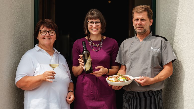 Drei Personen vor einem Restaurant, eine Frau mit Weinglas, eine Frau mit Weinflasche, ein Mann im Kochoutfit mit Teller.