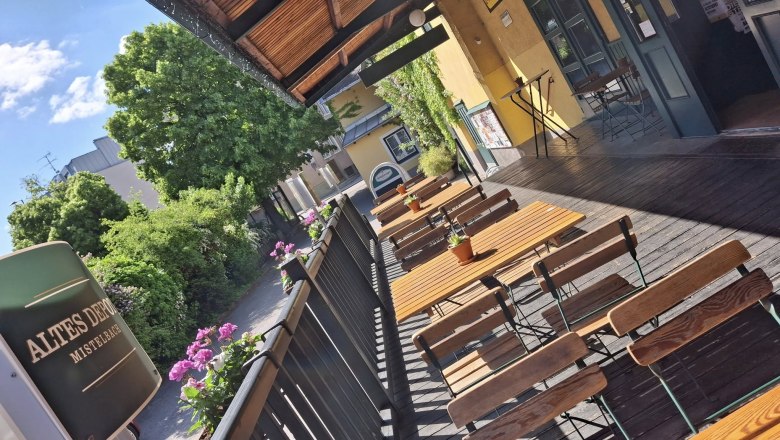 Eine sonnige Terrasse mit Holztischen und -st&uuml;hlen, umgeben von Pflanzen und einem Geb&auml;ude mit gelber Fassade.