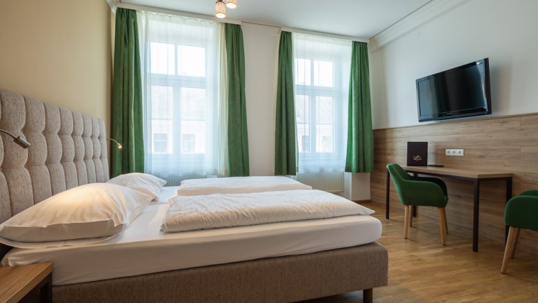 Hotelzimmer mit Doppelbett, grüne Vorhänge, Schreibtisch, Stuhl und Wand-TV.