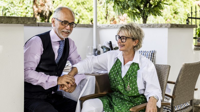 Ein Mann und eine Frau sitzen l&auml;chelnd an einem Tisch im Freien, sie halten sich an den H&auml;nden.