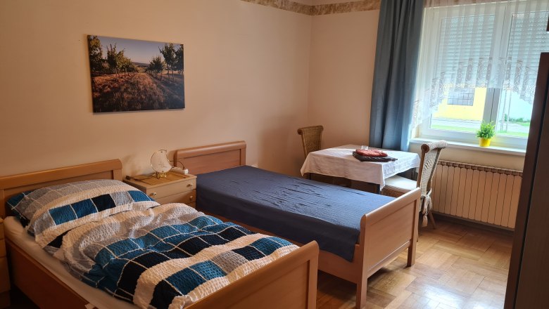 A simply furnished bedroom with two single beds, a table and chair and a window with curtains.