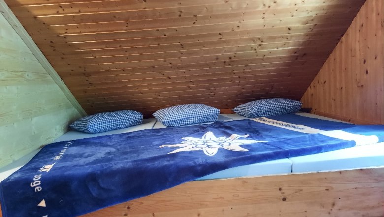 Matracov&aacute; ložnice s modrou dekou a kostkovan&yacute;mi pol&scaron;t&aacute;ři pod dřevěn&yacute;m stropem.