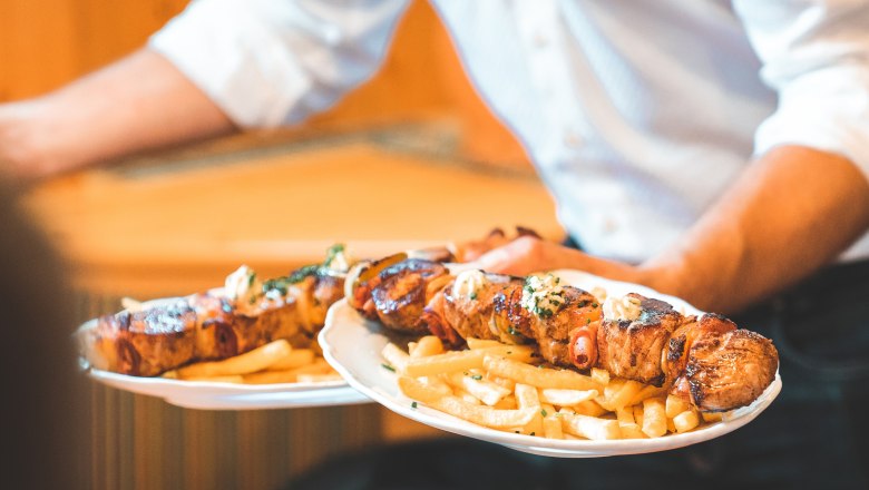 Ein Kellner serviert zwei Teller mit gegrillten Fleischspießen und Pommes frites.