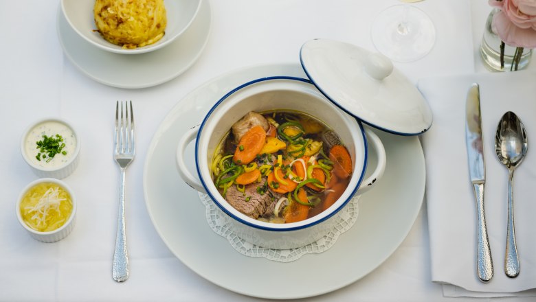 Ein gedeckter Tisch mit einem Eintopf in einem weißen Topf, einem Knödel, zwei Soßen und einem Glas Weißwein.