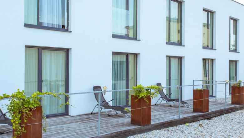 Moderne Terrasse mit Liegestühlen und Pflanzen vor einem Gebäude mit großen Fenstern.