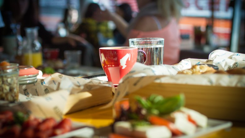 Ein roter Kaffeebecher und ein Glas Wasser auf einem Tisch in einem Caf&eacute;, umgeben von Essen und Getr&auml;nken.