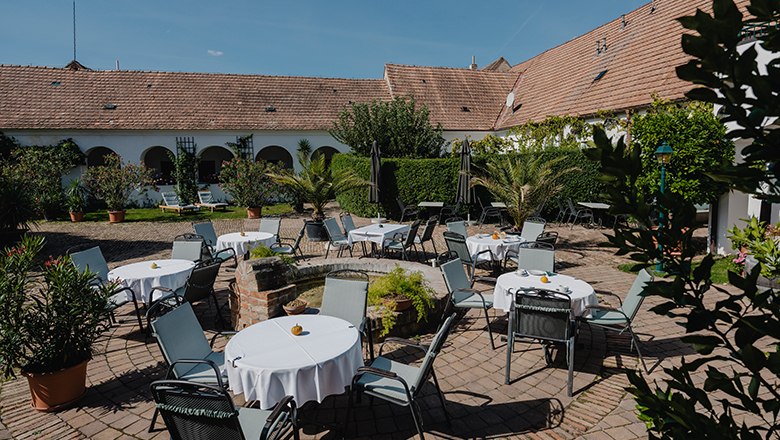 Ein gemütlicher Garten mit runden Tischen und Stühlen, umgeben von Pflanzen und einem alten Gebäude mit Ziegeldach.