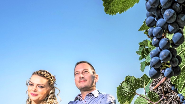 Ein Mann und eine Frau stehen vor einem blauen Himmel neben einer Weinrebe mit Trauben.
