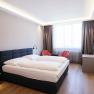 Modernes Hotelzimmer mit Doppelbett, zwei roten Sesseln, einem gro&szlig;en Fenster und einem Flachbildfernseher an der Wand.