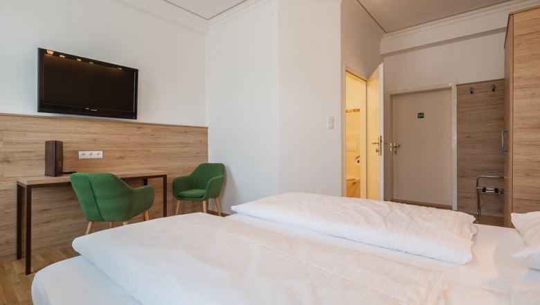 Hotelzimmer mit Doppelbett, grüne Stühle, Schreibtisch und Fernseher.