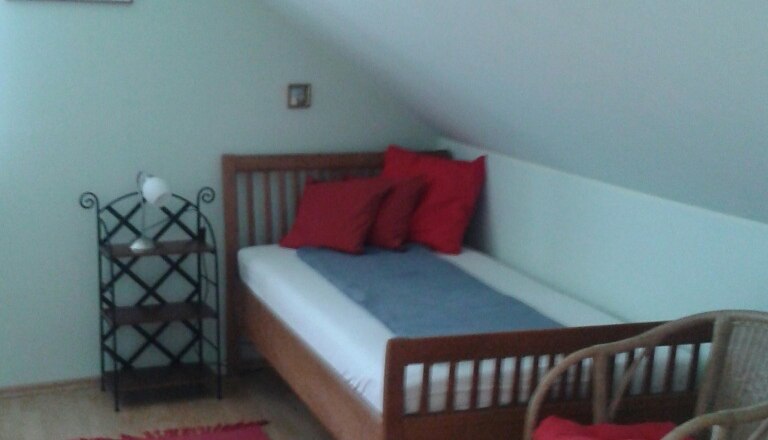 Ein Einzelbett in einem Zimmer mit Dachschr&auml;ge, rotem Teppich, Rattanstuhl und Regal.