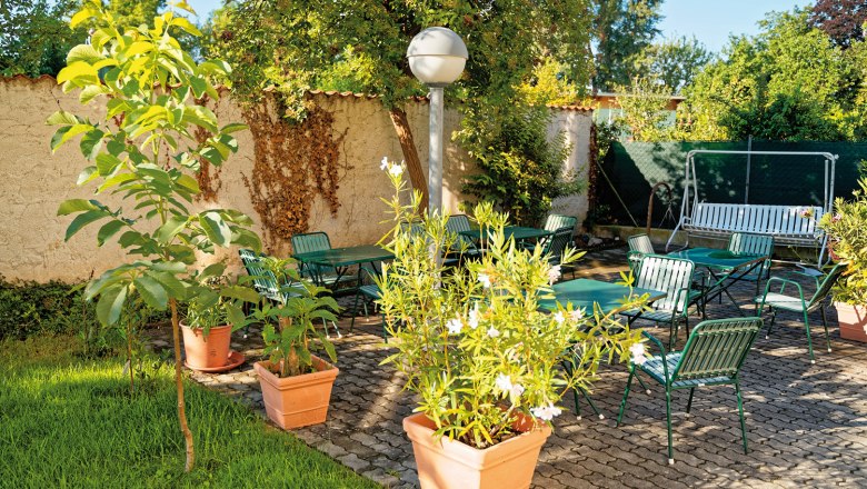 A sunny garden with green chairs, tables and plants in pots on a paved terrace.