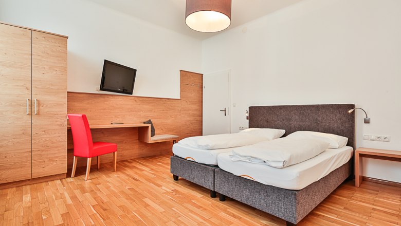 Hotelzimmer mit Doppelbett, rotem Stuhl, Schreibtisch und Fernseher.