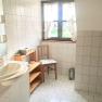 Helles Badezimmer mit Waschbecken, Holzregal, Stuhl und Fenster mit Vorhängen.
