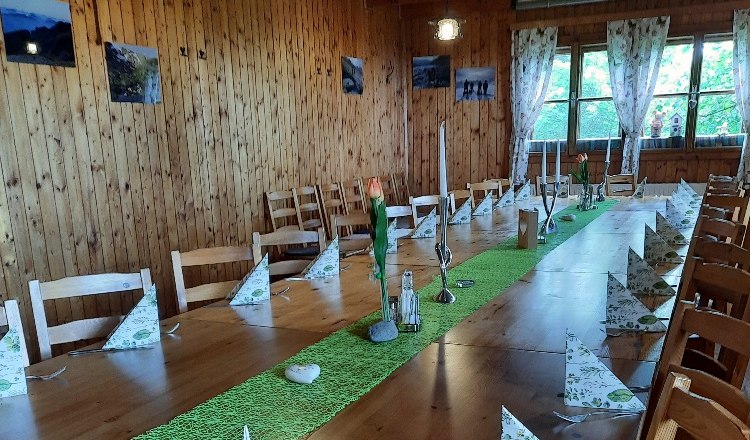 Ein rustikaler Raum mit Holzwänden und -decke. Ein langer Tisch ist mit grüner Tischdecke, Kerzen und gefalteten Servietten gedeckt.