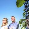 Ein Mann und eine Frau stehen vor einem blauen Himmel neben einer Weinrebe mit Trauben.