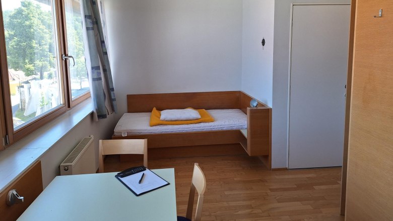 Jednodu&scaron;e zař&iacute;zen&yacute; jednolůžkov&yacute; pokoj s postel&iacute;, stolem a židl&iacute;, světl&yacute;m dřevěn&yacute;m n&aacute;bytkem a velk&yacute;m oknem s v&yacute;hledem ven.