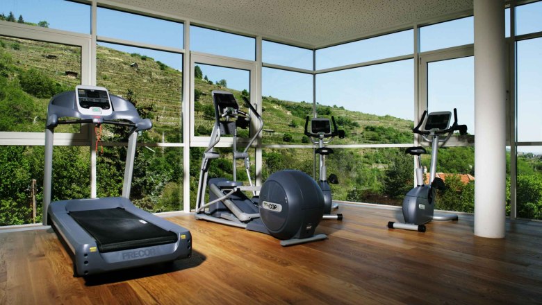 Fitnessraum mit Laufband, Crosstrainer und Heimtrainer vor großen Fenstern mit Blick auf grüne Hügel.