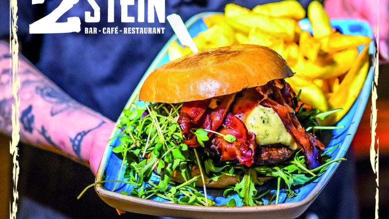 Ein Burger mit Speck, K&auml;se und Rucola auf einem Teller mit Pommes, pr&auml;sentiert von einer t&auml;towierten Hand. Logo von 2Stein Bar-Caf&eacute;-Restaurant oben links.