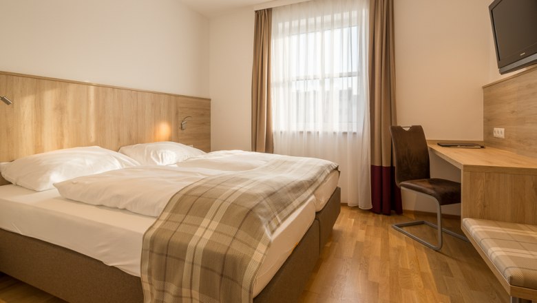 Hotelzimmer mit Doppelbett, Schreibtisch und Fenster mit Vorhängen.